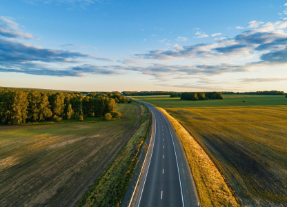 A winding road stretches into the distance, flanked by vibrant green fields and dense trees under a clear blue sky.